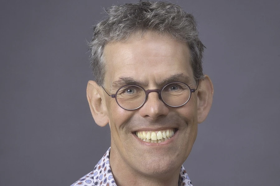 main with grey black hair in format shirt smiling against grey wall